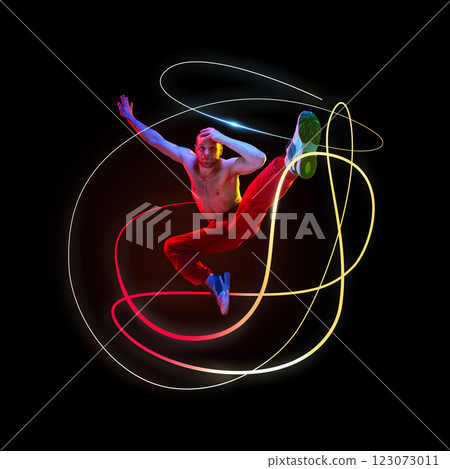 Dynamic image of shirtless man, dancer in motion performing highlighted by red and yellow neon light trails against dark space. Dynamic image of shirtless man, dancer in motion performing highlighted by red and yellow neon light trails against dark space. 123073011