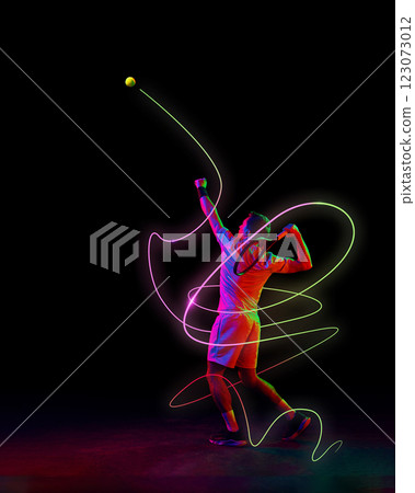 Focused man, tennis player in motion, serving ball with racket with neon green and pink light trails emphasizing motion against black background. Focused man, tennis player in motion, serving ball with racket with neon green and pink light trails emphasizing motion against black background. 123073012