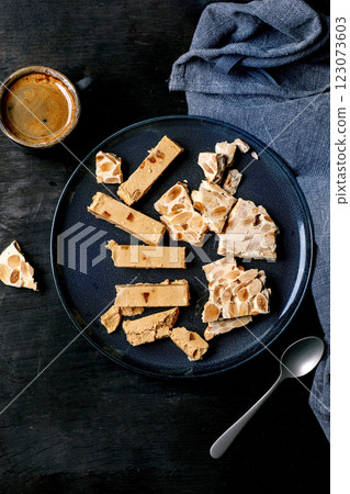 Traditional Spanish dessert classic soft orange turron and crunchy almond turron, cut into pieces on blue ceramic plate. Linen napkin, espresso coffee in black cup, black wooden background, top view. 123073603