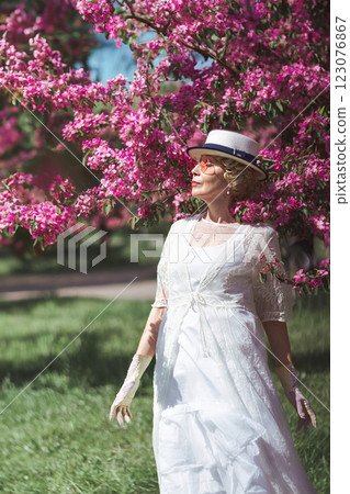 Happy natural adult mature woman of 60 years old in 18th century style white canotier hat, retro dress, gloves and glasses in blossoming pink apple orchard on summer day 123076867