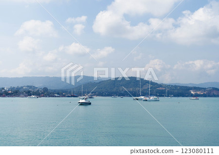 Calm waters reflect soft clouds as yachts float gently in a tranquil harbor, framed by rolling hills. This idyllic coastal setting invites relaxation and exploration under the midday sun. 123080111