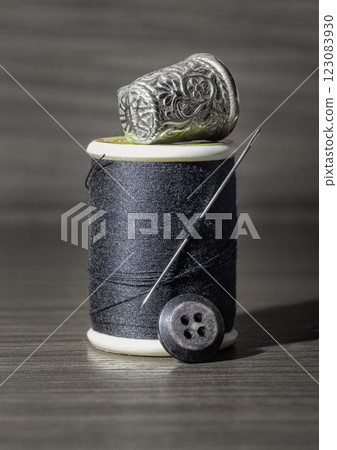 Silver sewing needle stuck in a spool of black thread with Silver thimble on top and black button in front of on wooden table. 123083930