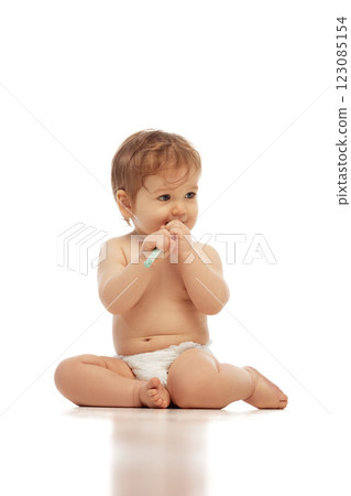 Little baby sitting, wearing in diaper, cross-legged while holding toothbrush and chewing on it against white background. 123085154