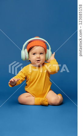Happy baby wearing bright orange beanie and headphones adjusting ear cups while playfully and funny dancing against deep blue background. Happy baby wearing bright orange beanie and headphones adjusting ear cups while playfully and funny dancing against deep blue background. 123085158