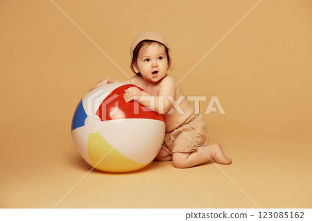 Portrait of playful little baby, wearing beige shorts and hat, hugging large colorful beach ball while kneeling against warm beige background. Portrait of playful little baby, wearing beige shorts and hat, hugging large colorful beach ball while kneeling against warm beige background. 123085162