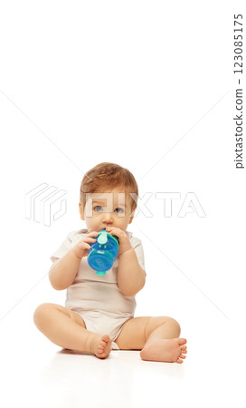 Little baby, dressed white onesie, sitting and drinking from blue sippy cup while holding it with both hands against white background. 123085175