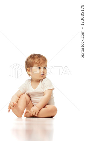 Baby, dressed white onesie, sitting cross-legged while looking sideways with focused expression against white background. Baby, dressed white onesie, sitting cross-legged while looking sideways with focused expression against white background. 123085179