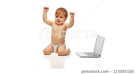 Happy baby, little explorer sitting and raising hands triumphantly next to open laptop against white background. 123085188