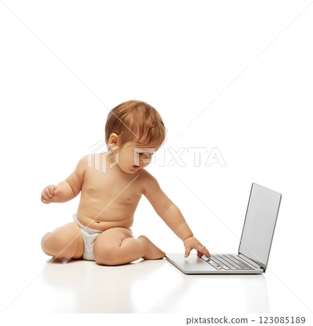 Portrait of little baby in diaper sitting and reaching out to touch laptop keyboard while looking at screen against white background. 123085189