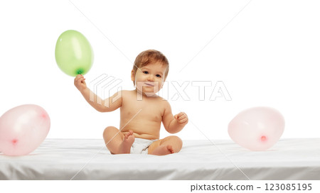 Little baby sitting on soft white bed, and holding green balloon while smiling, surrounded by pink balloons against white background. 123085195