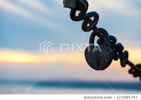 Close-up of a rusty closed heart-shaped padlock hanging on a metal chain on the seashore as a sign of love and loyalty for a wedding.. Padlock isolated on sky background. 123085742