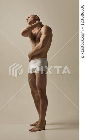 Shirtless male model dressed white underwear, tilting head with hand on neck in expressive movement against minimal studio background. 123086036