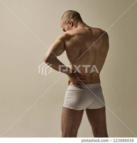 Back view of young, strong man holding lower back in poised stance with sculpted muscles against clean background. Back view of young, strong man holding lower back in poised stance with sculpted muscles against clean background. 123086038