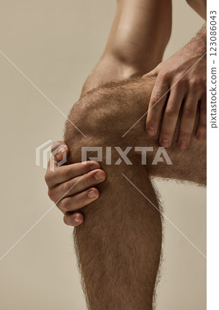 Close-up of strong male hands gripping knee in focused pose against warm studio background. 123086043