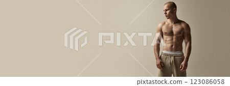 Banner. Athletic man standing with calm expression, gaze downward, muscular physique emphasized by soft lighting against neutral beige background. 123086058