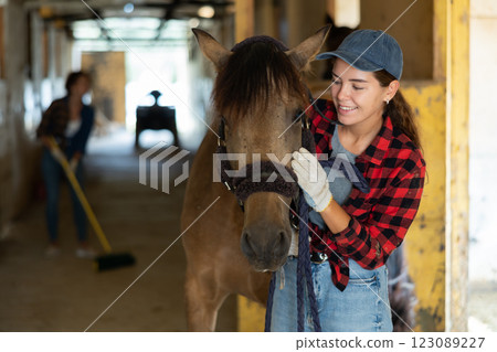 Young pretty girl farmer in plaid shirt and gloves petting and caressing horse, woman cleaning floor in background at stabling 123089227