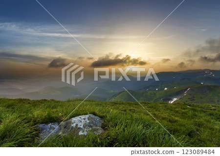 Wide summer mountain view at sunrise. Glowing orange sun raising in blue cloudy sky over green grassy hill with big rock and distant mountain range covered with morning mist. Beauty of nature concept. Wide summer mountain view at sunrise. Glowing orange sun raising in blue cloudy sky over green grassy hill with big rock and distant mountain range covered with morning mist. Beauty of nature concept. 123089484