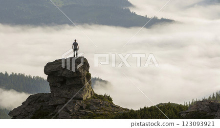 Silhouette of athletic climber tourist on high rocky formation on mountain valley filled with white puffy clouds and fog and covered with evergreen forest mountain slopes under clear sky background. 123093921