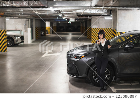 Confident Asian woman in 30s sending voice message in dimly lit urban parking. Professional attire suggests business setting as she leans against modern vehicle with phone in hand. Confident Asian woman in 30s sending voice message in dimly lit urban parking. Professional attire suggests business setting as she leans against modern vehicle with phone in hand. 123096897