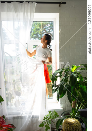 Woman hangs transparent tulle curtains on large windows in the house inside the interior with potted plants. Spring cleaning, tidying up Woman hangs transparent tulle curtains on large windows in the house inside the interior with potted plants. Spring cleaning, tidying up 123099093