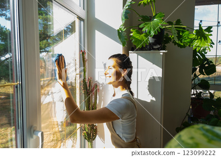 Woman in apron manually washes the window of house with rag cleaner and mop inside the interior with home plants on windowsill. Restoring order and cleanliness in the spring, cleaning servise Woman in apron manually washes the window of house with rag cleaner and mop inside the interior with home plants on windowsill. Restoring order and cleanliness in the spring, cleaning servise 123099222