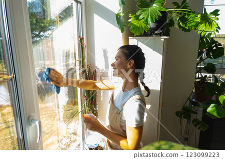 Woman in apron manually washes the window of house with rag cleaner and mop inside the interior with home plants on windowsill. Restoring order and cleanliness in the spring, cleaning servise Woman in apron manually washes the window of house with rag cleaner and mop inside the interior with home plants on windowsill. Restoring order and cleanliness in the spring, cleaning servise 123099223