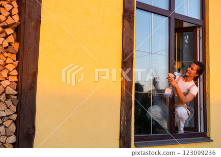 Woman in apron manually washes the window of the house with a rag with a spray cleaner and a mop outside. Safety at height, restoring order and cleanliness in the spring, cleaning service 123099236