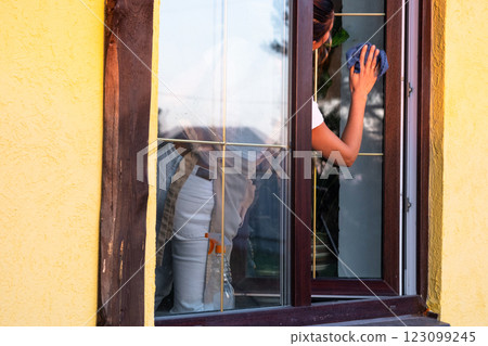 Woman in apron manually washes the window of the house with a rag with a spray cleaner and a mop outside. Safety at height, restoring order and cleanliness in the spring, cleaning service Woman in apron manually washes the window of the house with a rag with a spray cleaner and a mop outside. Safety at height, restoring order and cleanliness in the spring, cleaning service 123099245