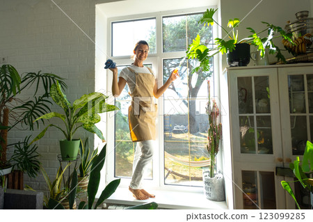 Woman Portrait in apron manually washes the window of house with rag cleaner and mop inside the interior with home plants on windowsill. Restoring order and cleanliness in the spring, cleaning servise Woman Portrait in apron manually washes the window of house with rag cleaner and mop inside the interior with home plants on windowsill. Restoring order and cleanliness in the spring, cleaning servise 123099285