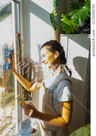 Woman in apron manually washes the window of house with rag cleaner and mop inside the interior with home plants on windowsill. Restoring order and cleanliness in the spring, cleaning servise Woman in apron manually washes the window of house with rag cleaner and mop inside the interior with home plants on windowsill. Restoring order and cleanliness in the spring, cleaning servise 123099289