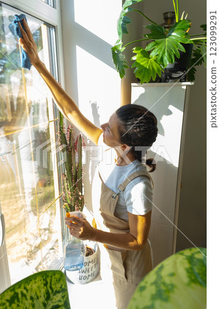 Woman in apron manually washes the window of house with rag cleaner and mop inside the interior with home plants on windowsill. Restoring order and cleanliness in the spring, cleaning servise Woman in apron manually washes the window of house with rag cleaner and mop inside the interior with home plants on windowsill. Restoring order and cleanliness in the spring, cleaning servise 123099291