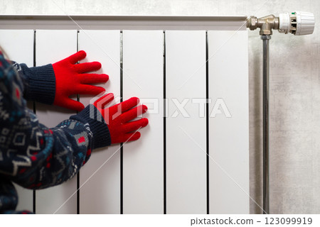 Hands in gloves on a radiator. Central heating radiator at home. A child in woolen gloves warms his hands near the heater Hands in gloves on a radiator. Central heating radiator at home. A child in woolen gloves warms his hands near the heater 123099919