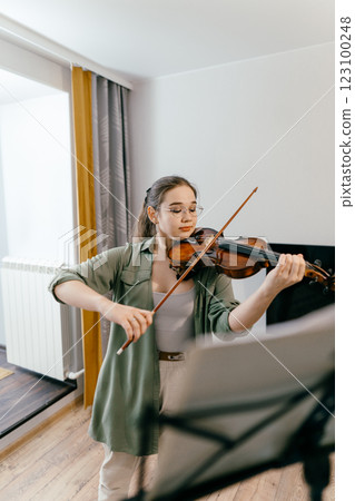 Young woman playing violin at home by sheet music, focused on learning music. Violin student rehearsing classical music in a professional study session 123100248