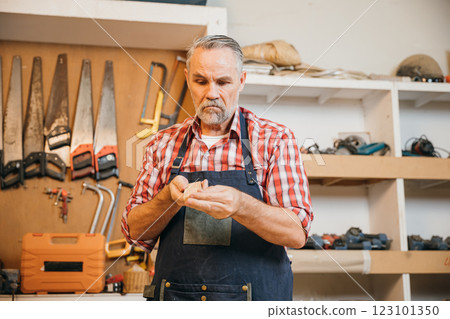 A senior carpenter in red plaid shirt carefully sands piece of wood in his workshop. Tools hang organized on wall behind him, illustrating his skill, dedication and attention to detail in woodworking A senior carpenter in red plaid shirt carefully sands piece of wood in his workshop. Tools hang organized on wall behind him, illustrating his skill, dedication and attention to detail in woodworking 123101350