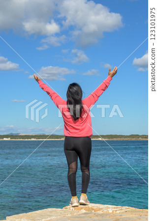 Woman facing the sea with arms raised, wearing pink and black, exuding joy under a blue sky, Woman facing the sea with arms raised, wearing pink and black, exuding joy under a blue sky, 123101975