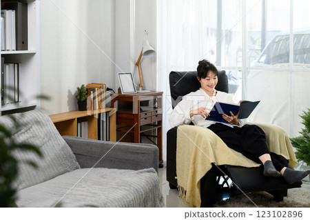 Pretty young woman sitting in a cozy armchair and reading a book with a smile Pretty young woman sitting in a cozy armchair and reading a book with a smile 123102896