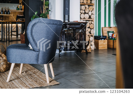 Cozy interior with a modern gray chair, woven basket, wood-burning stove, and stacked firewood, creating a warm and inviting atmosphere 123104263