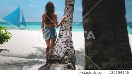 Young woman is taking a stroll on a pristine white sand beach, pausing by a palm tree with a traditional sailboat in the background 123104396