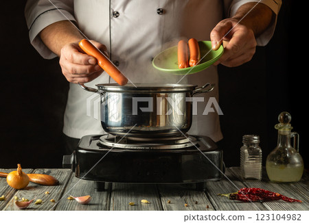 In a warm kitchen, a chef skillfully adds sausages to a pot, surrounded by fresh vegetables and spices, creating a delightful meal. The atmosphere is inviting and aromatic 123104982