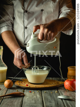A skilled chef uses a handheld mixer to blend milk and eggs in a bowl, surrounded by fresh apples and ingredients, evoking a warm and inviting culinary atmosphere A skilled chef uses a handheld mixer to blend milk and eggs in a bowl, surrounded by fresh apples and ingredients, evoking a warm and inviting culinary atmosphere 123104986