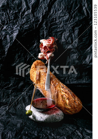 Still life with sliced Spanish ham Jamon Iberico, paleta on fork, glass of red wine with single olive, white bread loaf. Crumpled black paper background. Concept of traditional Spanish cuisine. 123105539