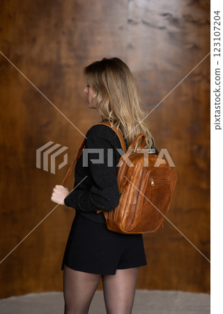 A girl in a warm sweater, a black short skirt and nylon tights is posing with a brown backpack. 123107204