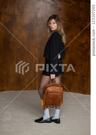 A girl in a warm sweater, a black short skirt, nylon tights, and black shoes is posing with a brown backpack. A girl in a warm sweater, a black short skirt, nylon tights, and black shoes is posing with a brown backpack. 123107205