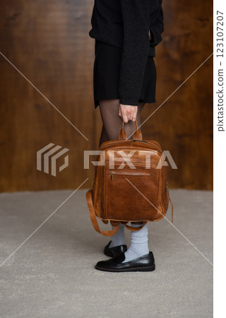 A girl in a warm sweater, a black short skirt, nylon tights, and black shoes is posing with a brown backpack. A girl in a warm sweater, a black short skirt, nylon tights, and black shoes is posing with a brown backpack. 123107207