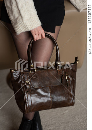 A girl in a warm sweater, a black short skirt, nylon tights, and black shoes is posing with a brown leather bag. 123107335