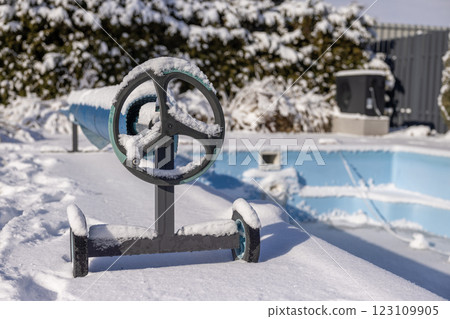 A snow-covered pool cover roller next to a frozen swimming pool in a backyard, surrounded by winter scenery with frost-covered trees and outdoor equipment. A snow-covered pool cover roller next to a frozen swimming pool in a backyard, surrounded by winter scenery with frost-covered trees and outdoor equipment. 123109905