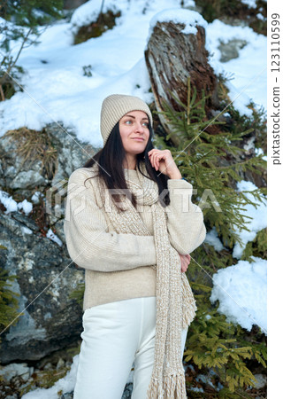 Young woman in cream-colored knitted sweater and beanie standing in a serene snowy mountain landscape with rocky outcrops and evergreen trees, exuding a contemplative and wintery atmosphere. 123110599
