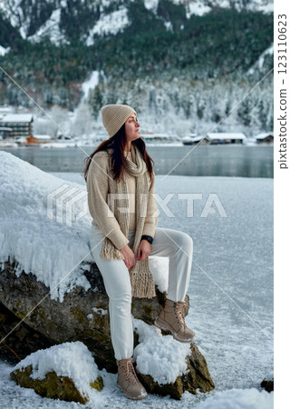 Young woman in cozy winter attire sitting on snowy rock with serene Eibsee lake and Zugspitze mountain view in background, exuding peaceful winter atmosphere. Young woman in cozy winter attire sitting on snowy rock with serene Eibsee lake and Zugspitze mountain view in background, exuding peaceful winter atmosphere. 123110623