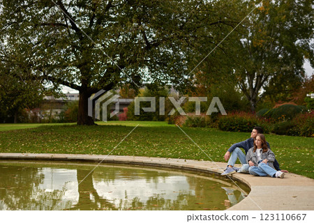 Autumn Serenity: Couple Relaxing by Reflective Pond Amidst Vibrant Fall Foliage and Ivy-Covered Architecture. Tranquil Garden Scene Captures Intimate Moment Against Backdrop of Colorful Autumn Leaves 123110667