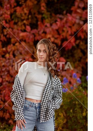 Young woman in casual autumn outfit posing against vibrant red ivy wall, stylish fashion portrait in warm seasonal tones, cozy fall atmosphere, urban outdoor setting, trendy seasonal style Young woman Young woman in casual autumn outfit posing against vibrant red ivy wall, stylish fashion portrait in warm seasonal tones, cozy fall atmosphere, urban outdoor setting, trendy seasonal style Young woman 123110669